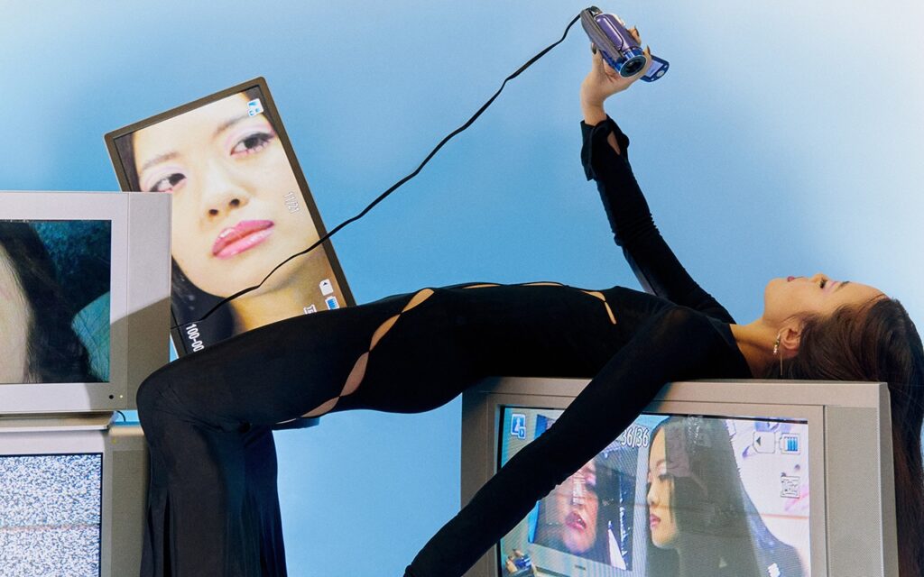 A girl laying on a retro tv holding a camcorder that is connected to tv and you can see her face in the screens